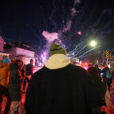 People on a street at night react as small fireworks explode in the air, with flashing emergency lights in the background.