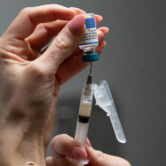 A close-up shows a person's hands holding a syringe and an inverted vaccine vial while drawing liquid medication into the syringe.