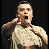 Comedian Carlos Mencia points toward the audience while speaking into a microphone during a stand-up performance.