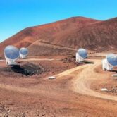 Several white radio telescope dishes sit across a barren volcanic landscape beneath a clear blue sky, with reddish-brown hills rising in the background.